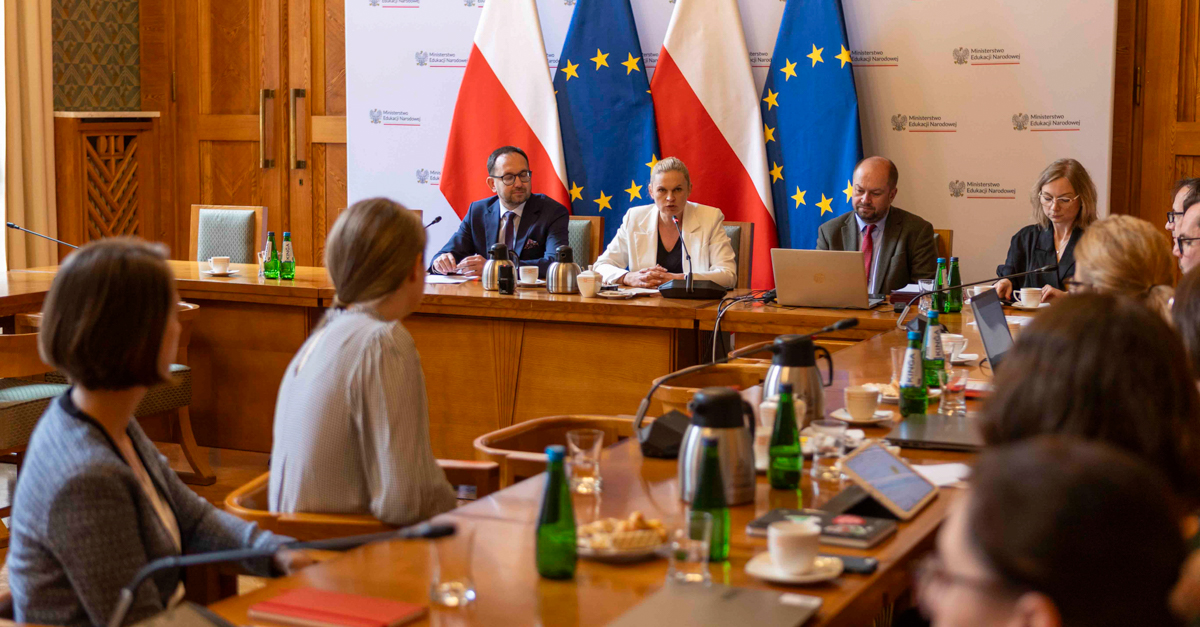Zdjęcie przedstawiające grupę ludzi w sali podczas posiedzenia, trzy osoby siedzą na tle flag Polski oraz UE.